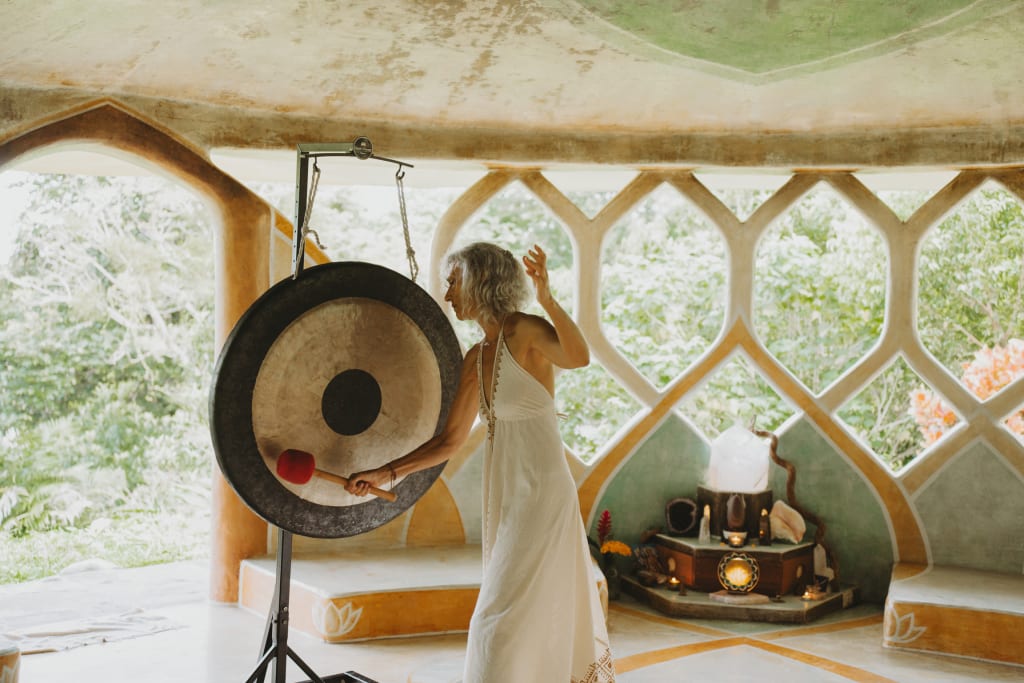 A person in a white dress is striking a large gong with a mallet in a room with arched windows. The space has a serene atmosphere with nature visible outside. An altar with candles and a crystal is in the background.